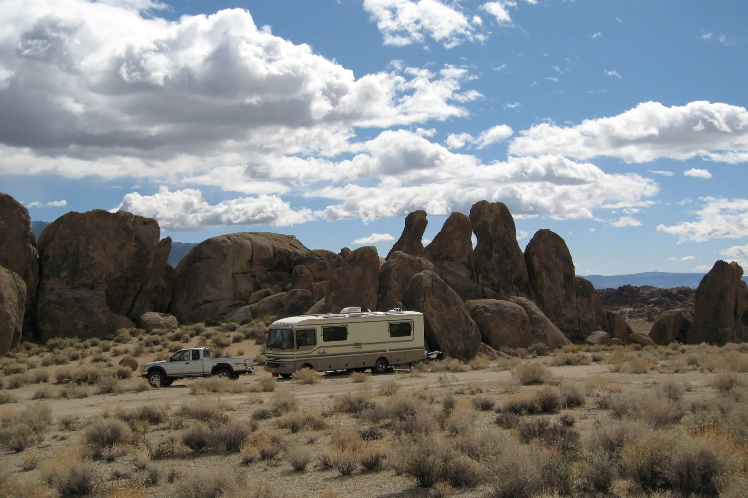 An older Fleetwood Bounder and a pickup truck parked among granite boulders and sagebrush under a cloudy blue sky.